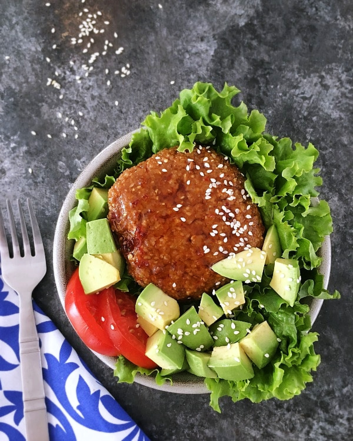 Overhead view of a ginger sesame teriyaki burger sitting on a bed of curly lettuce, tomato slices, and cubed avocado. The whole dish is topped with white sesame seeds, and a fork on a blue and white napkin sits alongside.