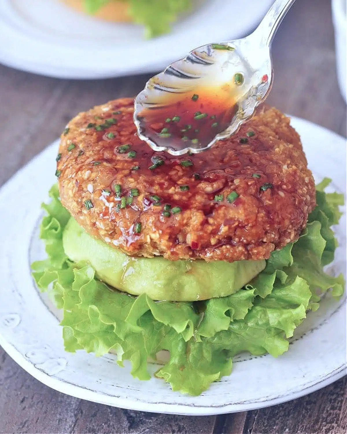 A ginger sesame teriyaki burger sitting on a bottom bun with lettuce and a thick slice of avocado, with more teriyaki sauce being spooned on top of the burger.