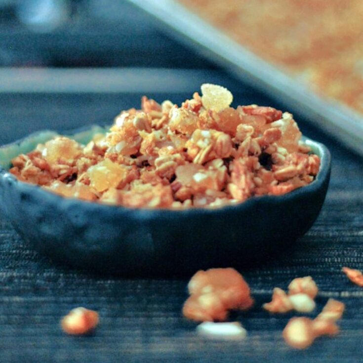 Golden colored ginger vanilla granola in a small black handmade bowl, with a few granola pieces on the tabletop. Full baking sheet of granola blurred in background.