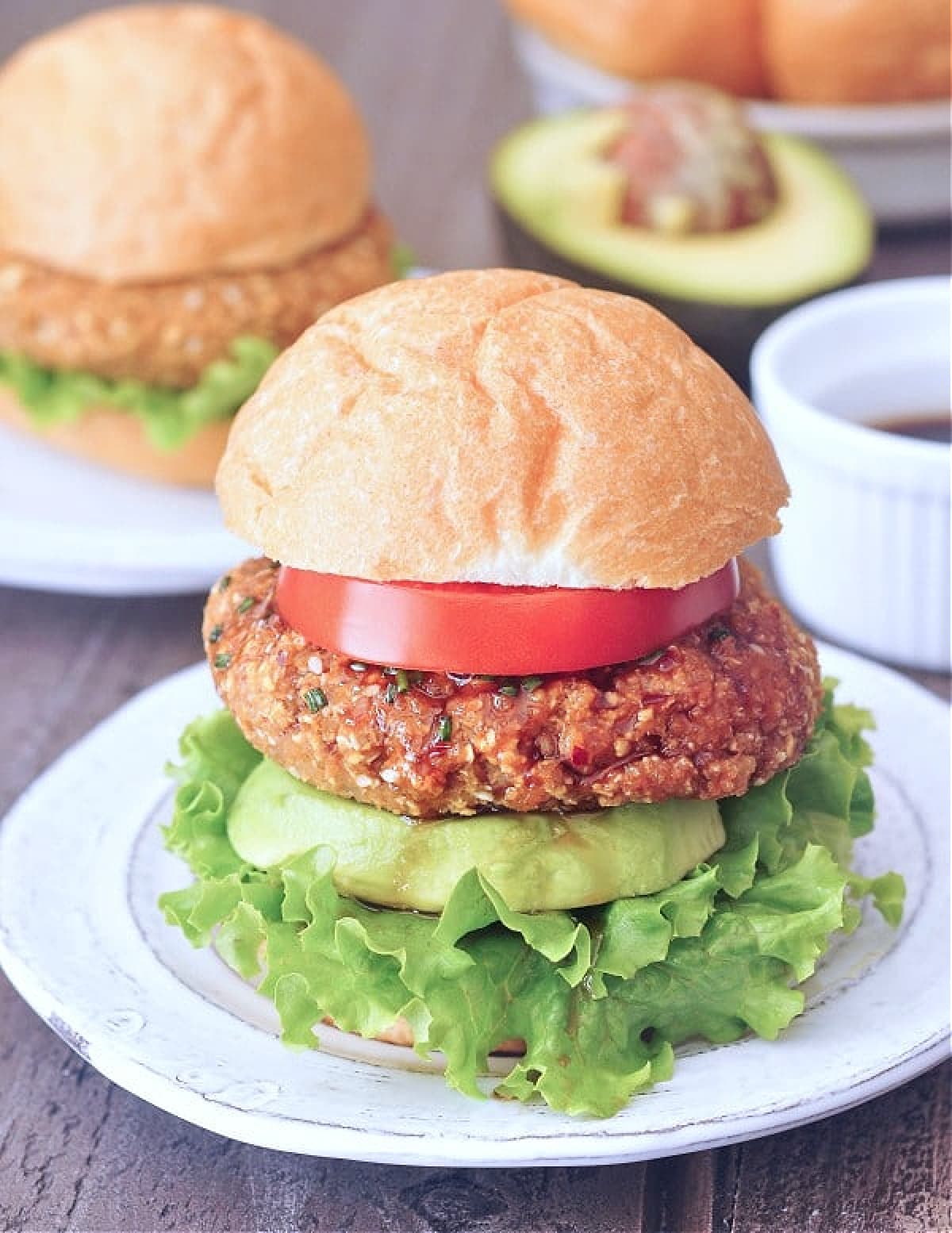 A ginger sesame teriyaki burger sitting on a bottom bun with lettuce, a thick slice of avocado, bright red slice of tomato, and top bun. More burgers, buns, and avocado in background.