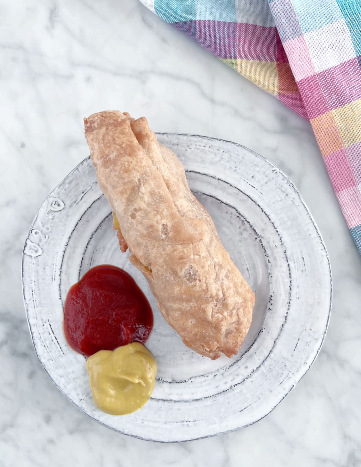 Overhead view of a hot dog with cheese wrapped in baked pastry sitting on a rustic grey dish with blobs of bright red ketchup and classic yellow mustard. Sitting on a white marble table with a pastel plaid cloth napkin.