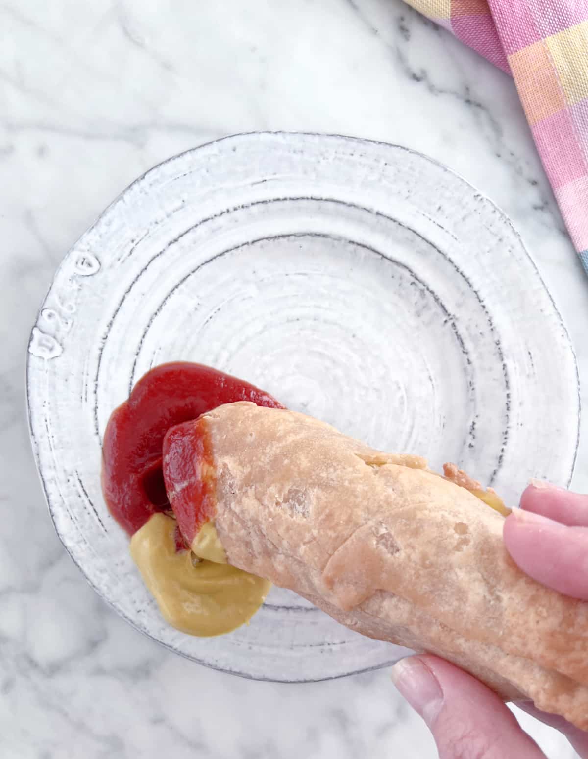 A hand dipping a wiener wrap (a hot dog with cheese wrapped in pastry and baked) in ketchup and mustard. Sitting on a white marble table with a pastel plaid cloth napkin.