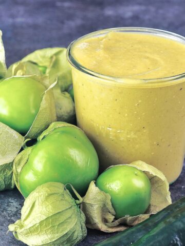 Close up of homemade green tomatillo enchilada sauce in a glass canning jar, surrounded by whole fresh tomatillos and peppers.