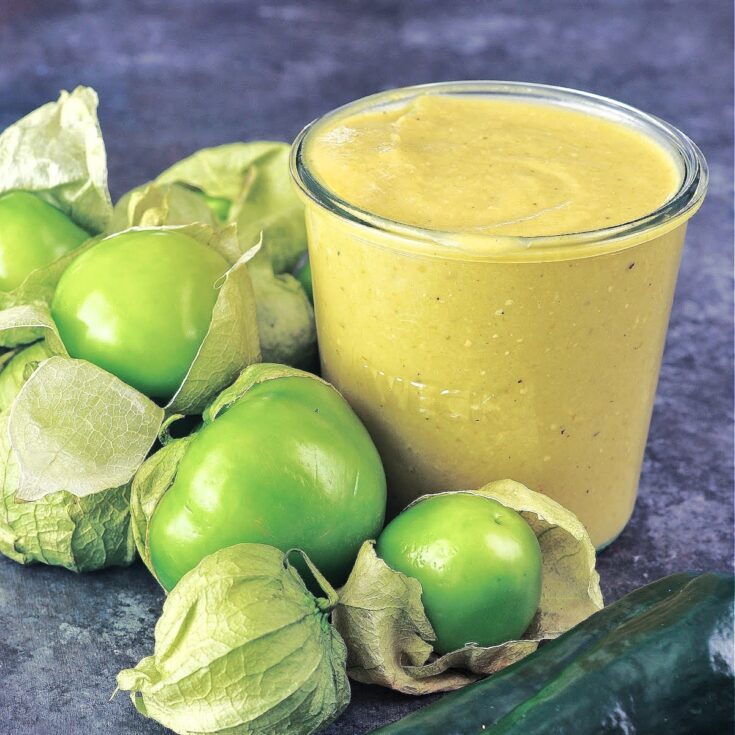 Close up of homemade green tomatillo enchilada sauce in a glass canning jar, surrounded by whole fresh tomatillos and peppers.