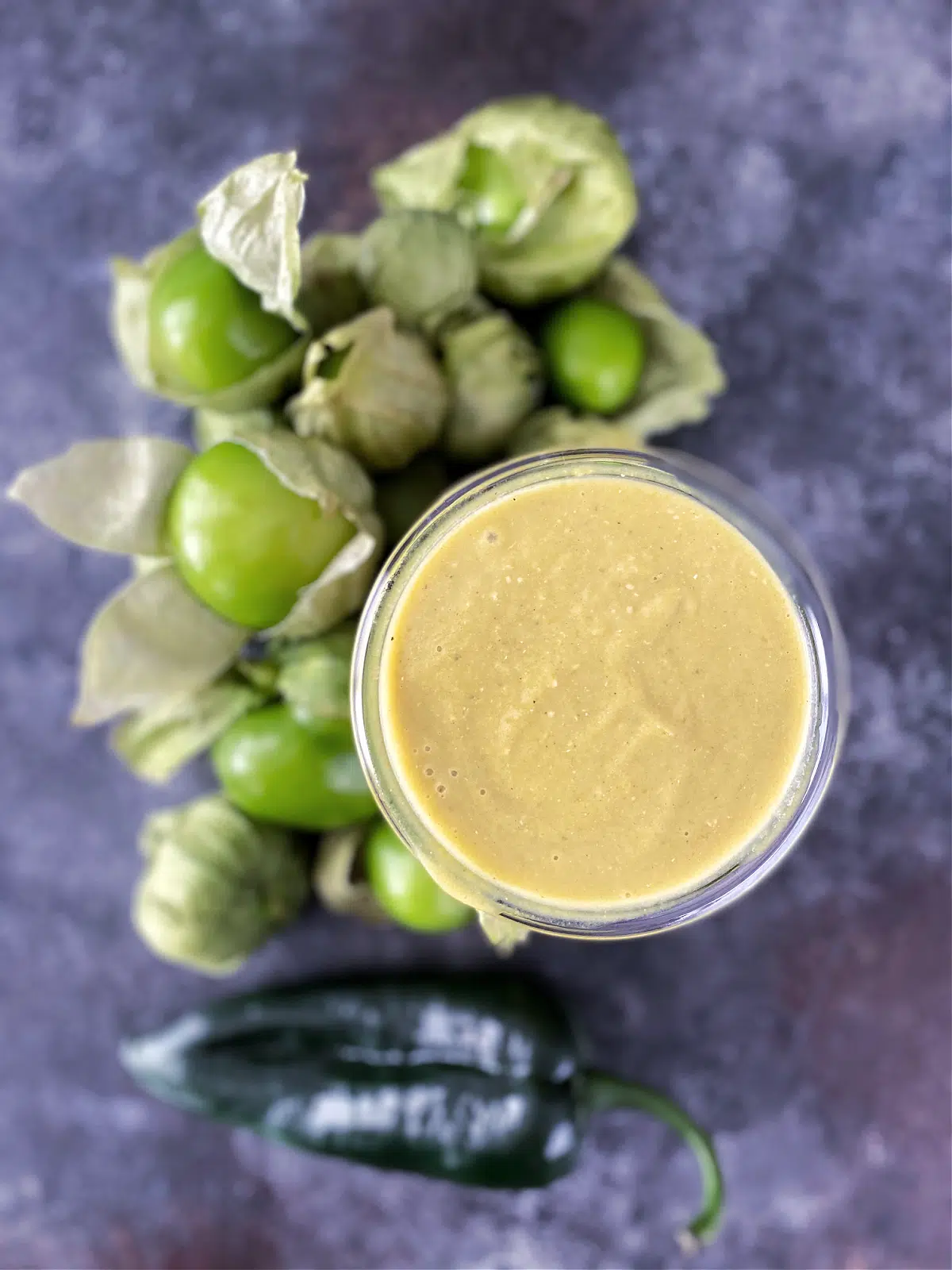 Overhead view of homemade green tomatillo enchilada sauce in a glass canning jar, surrounded by whole fresh tomatillos and peppers.