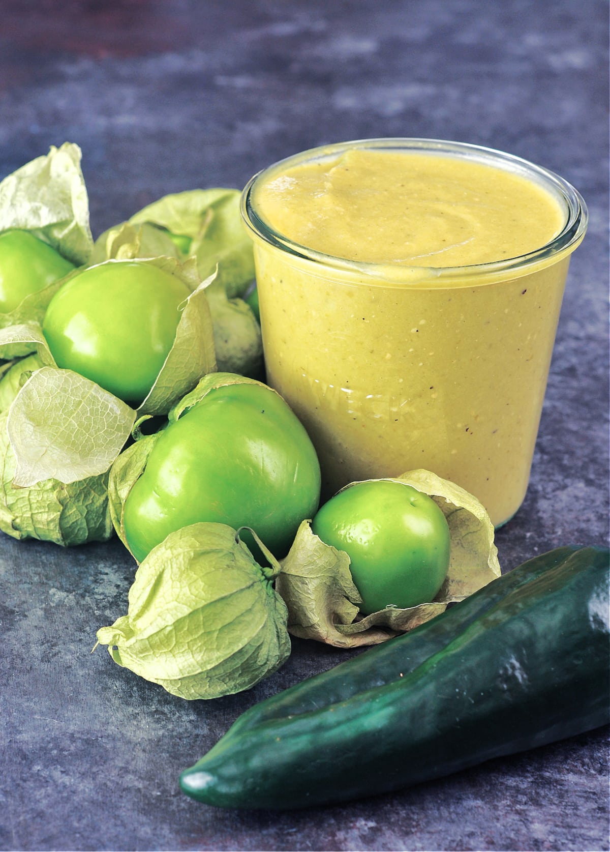 Homemade green tomatillo enchilada sauce in a glass canning jar, surrounded by whole fresh tomatillos and peppers.