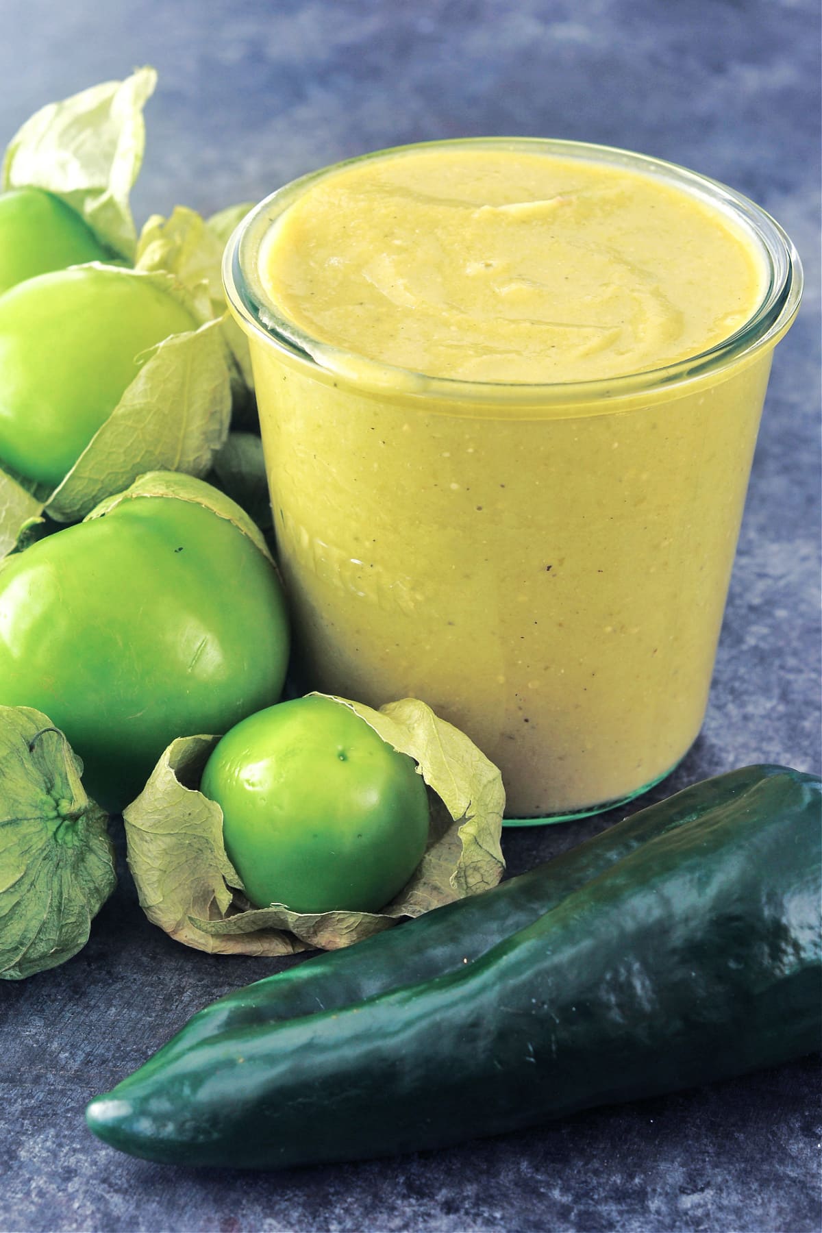 Homemade green enchilada sauce in a glass canning jar, surrounded by whole fresh tomatillos and peppers.
