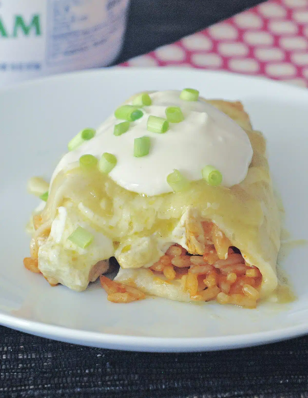 One cheesy Hatch chile enchilada covered in green enchilada sauce, sour cream, and diced green onion on a white plate, a tub of sour cream in background.