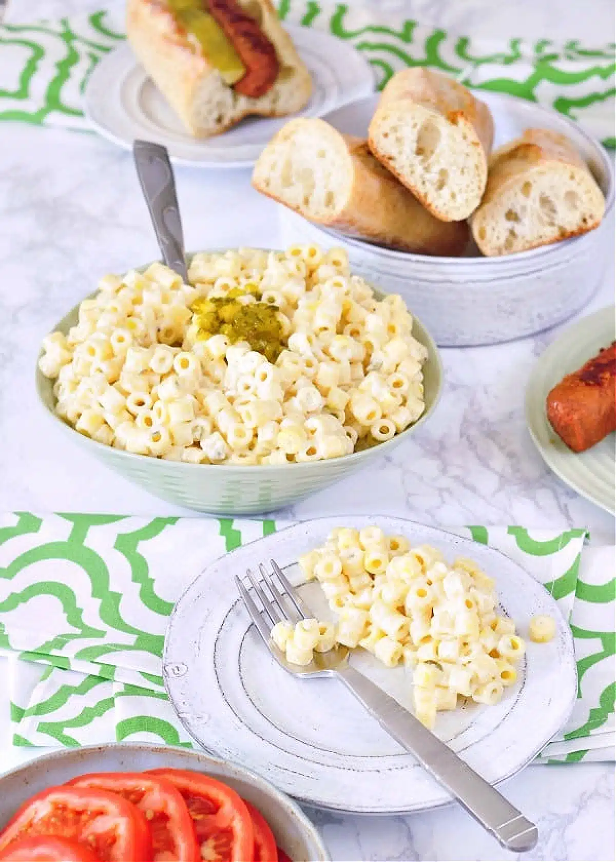 Hawaiian mac salad in a green serving bowl, with a smaller plate of mac salad on the side. More plates with hot dogs and a dish of hot dog rolls also on table.