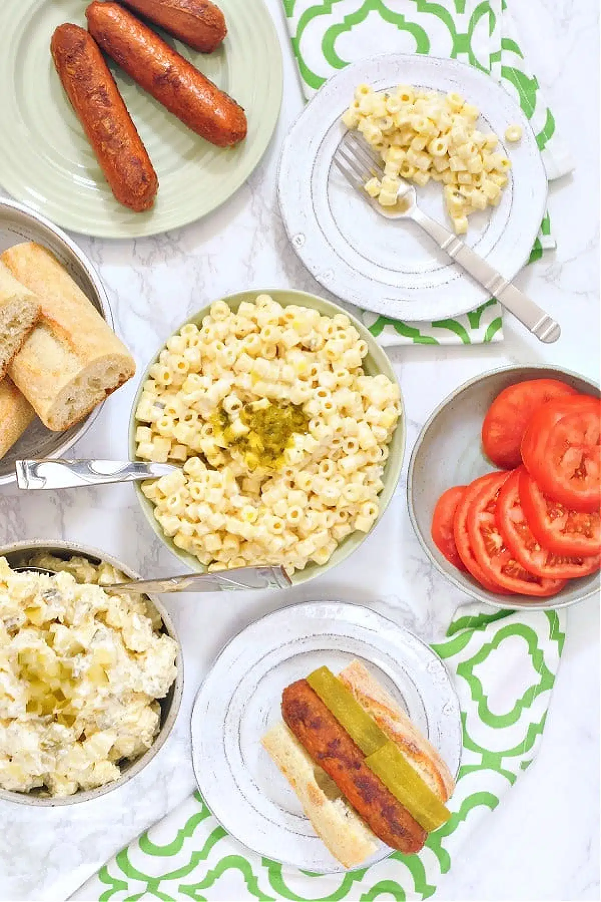 Overhead view of a picnic scene: Hawaiian mac salad in a green serving bowl, with a smaller plate of mac salad on the side. More plates with hot dogs, a plate of sliced tomatoes, and a dish of hot dog rolls also on table.