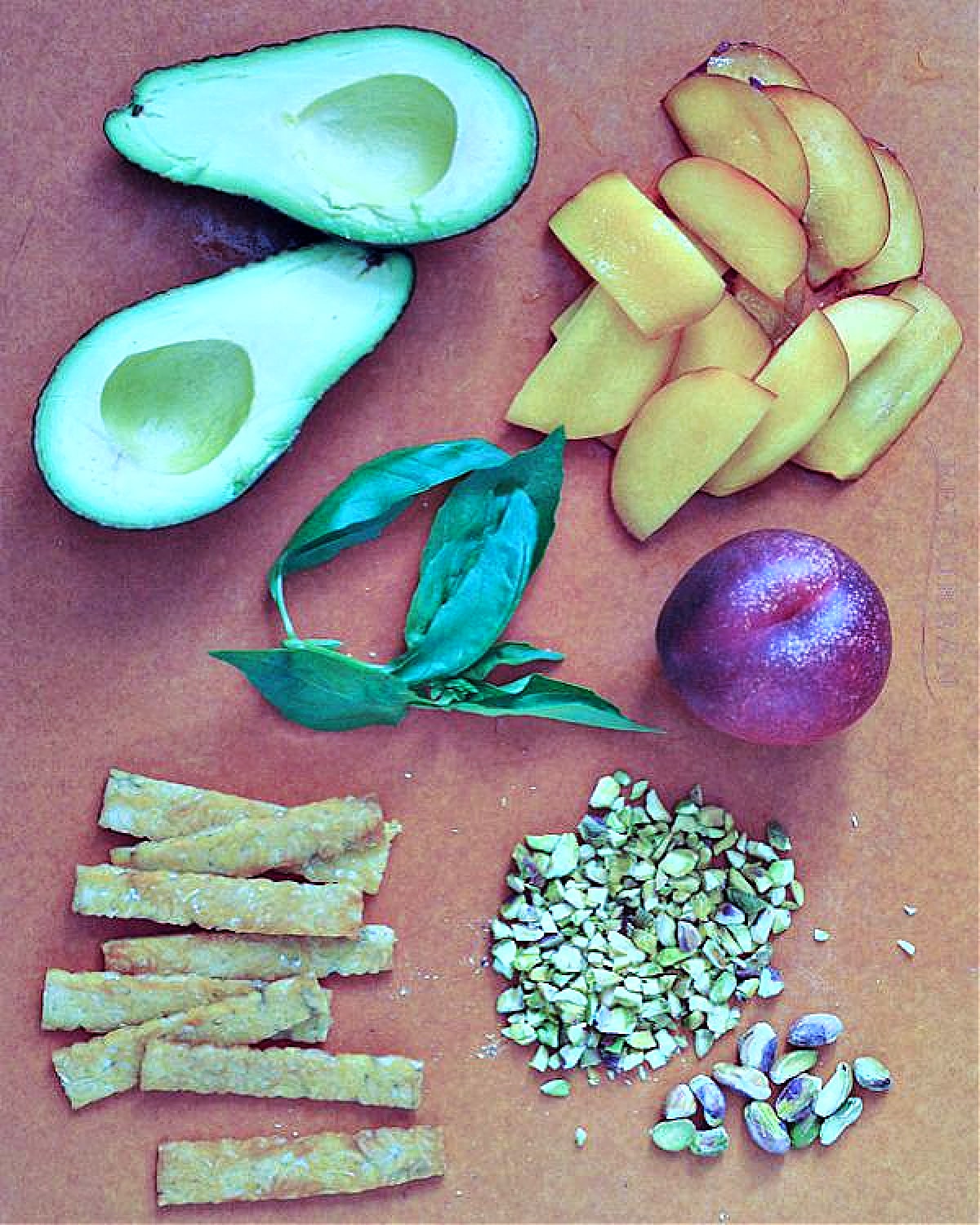 Ingredients for summer rolls on a wood cutting board: pan fried tempeh, sliced nectarines, sliced avocado, fresh basil leaves, pistachios, rice paper.