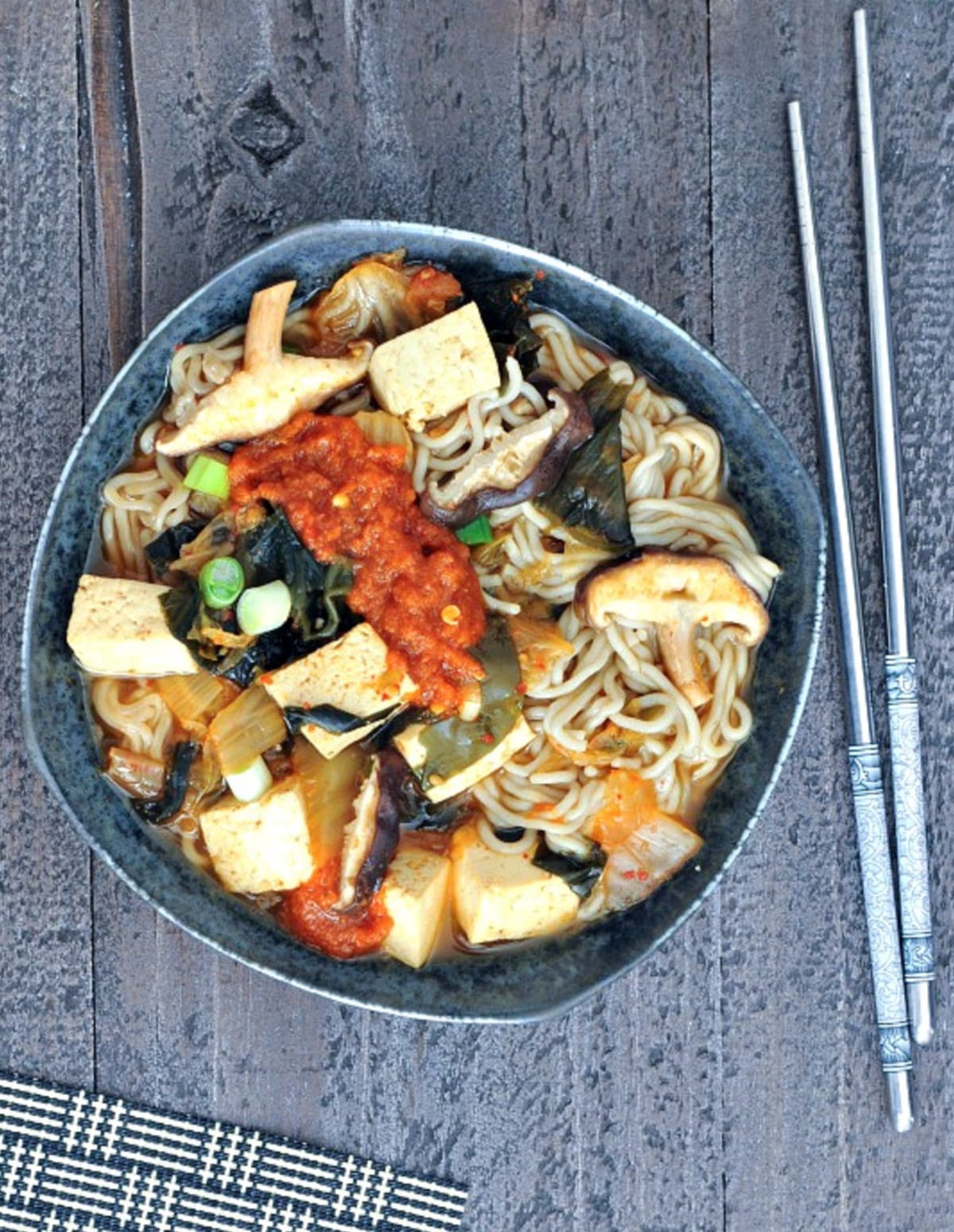 Overhead view of a bowl of colorful instant kimchi noodle soup with tofu, mushrooms, broth, noodles, kimchi, scallions.