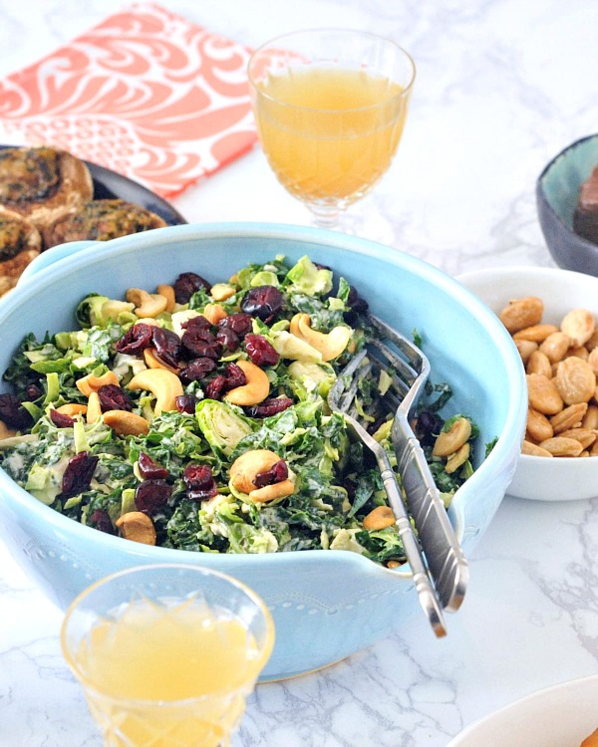 Lemon brussels and kale salad with cashews and dried cranberries in a light blue bowl, with glasses of champagne, nuts, and an orange napkin.