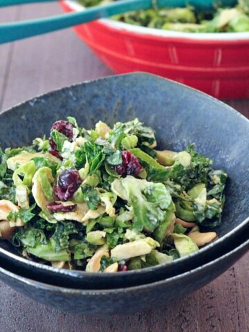 Lemon brussels and kale salad with cashews and dried cranberries in a dark grey bowl, with the large red serving bowl of salad in background.