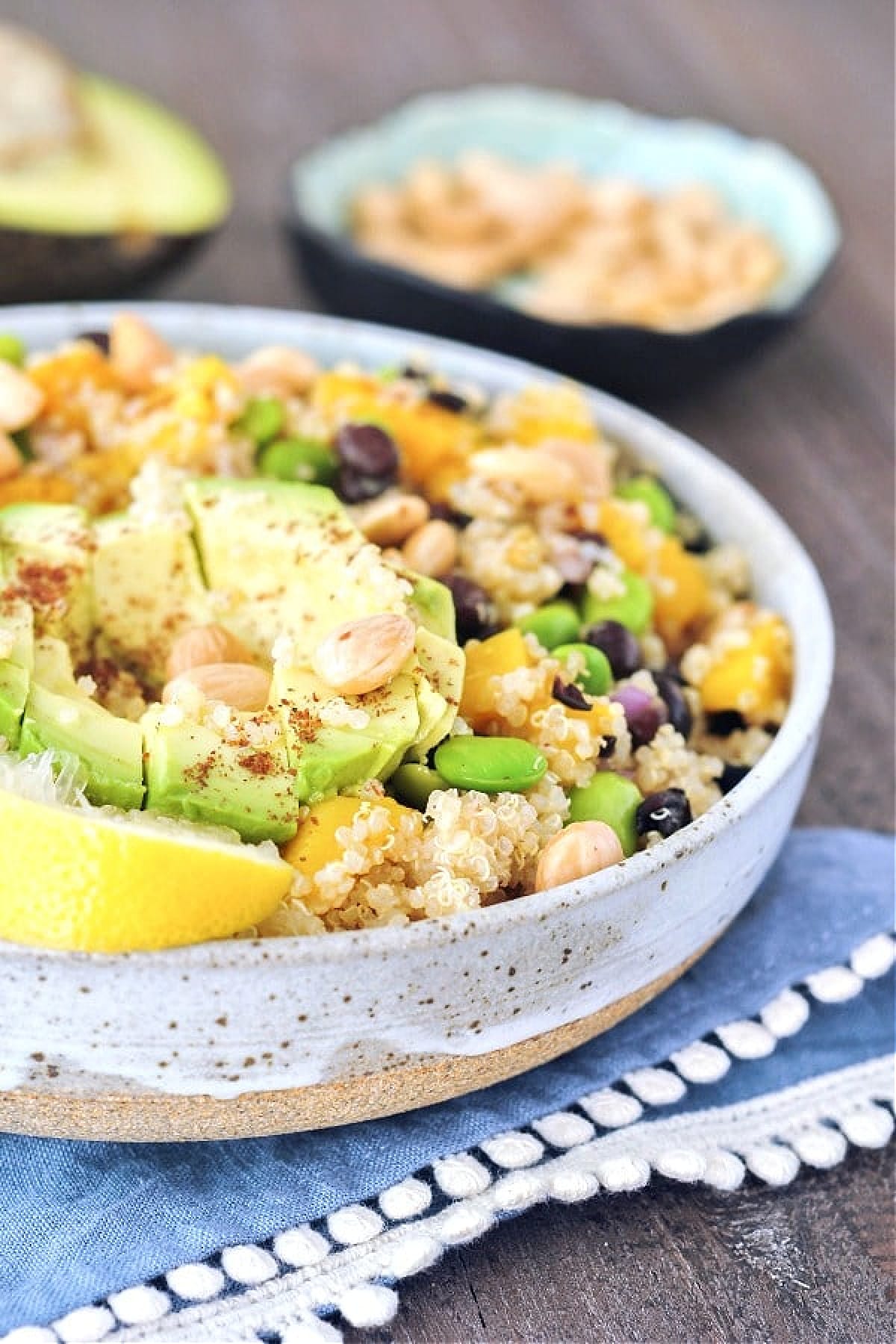 Mango edamame quinoa salad with cubed avocado on top. Image shows three quarters of the bowl, blurred background has a small bowl of almonds and another half avocado.