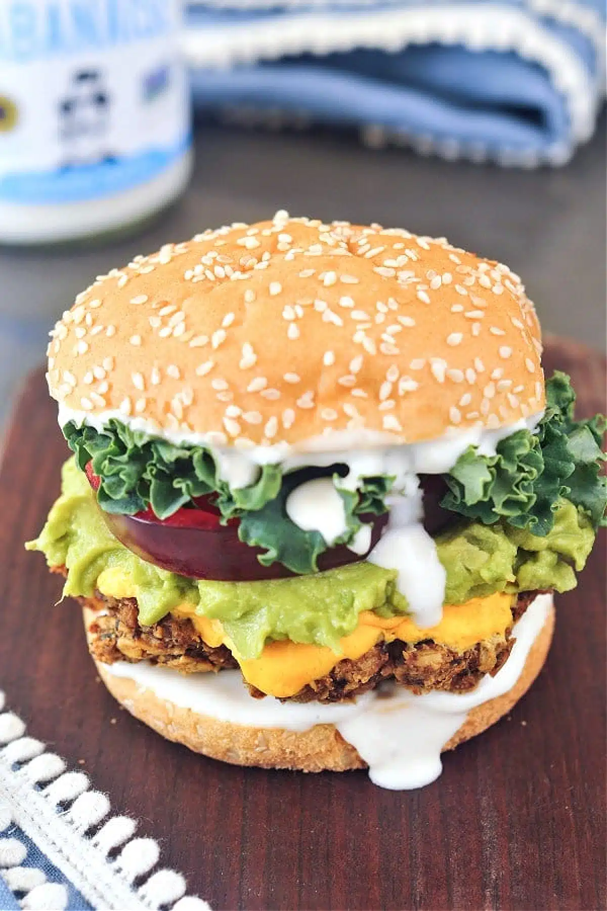 Mushroom burger sitting on a dark wood cutting board, mayo dripping down side. Colorful toppings include green curly lettuce, deep red thick tomato slice, light green mashed avocado, bright yellow vegan cheese sauce, bright white vegan mayonnaise. Mayonnaise jar and blue napkin blurred in background.