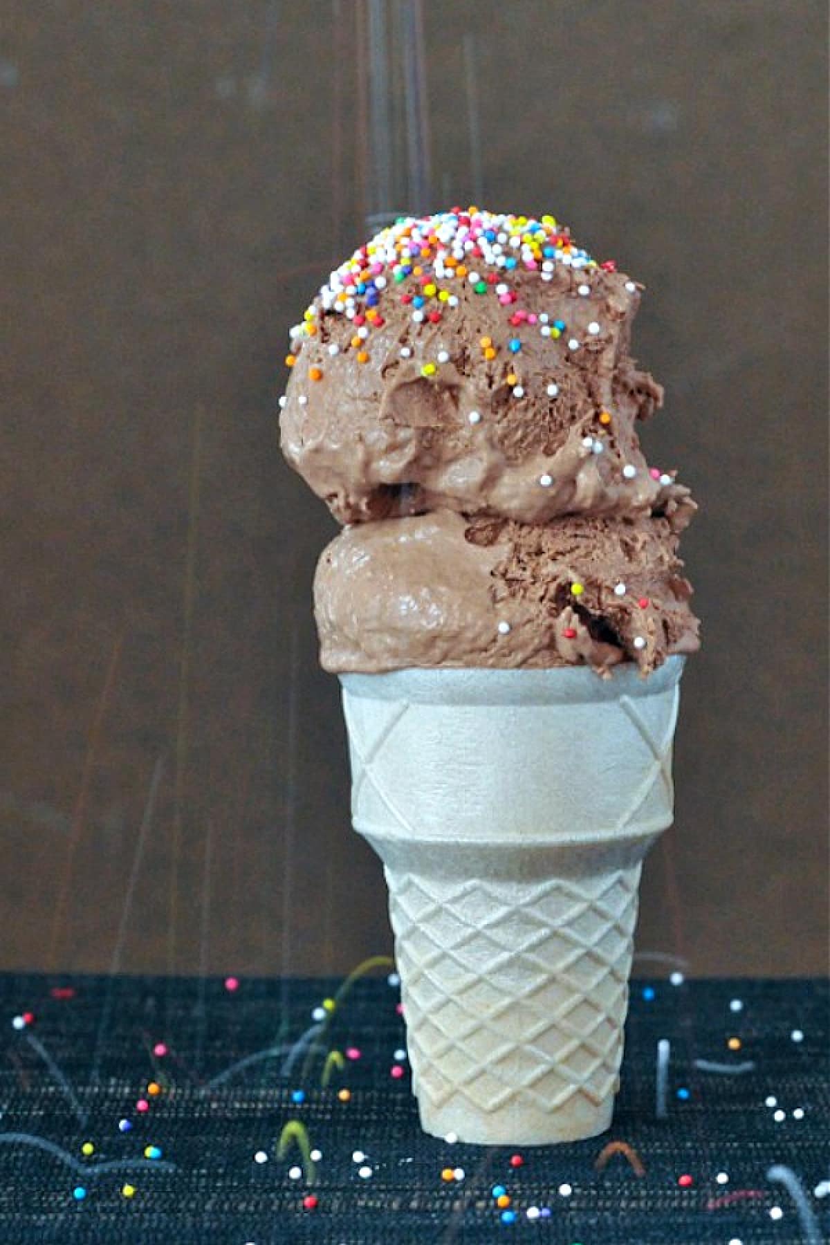 Two scoops of chocolate oat milk ice cream with rainbow colored sprinkles in a cone against a brown background.