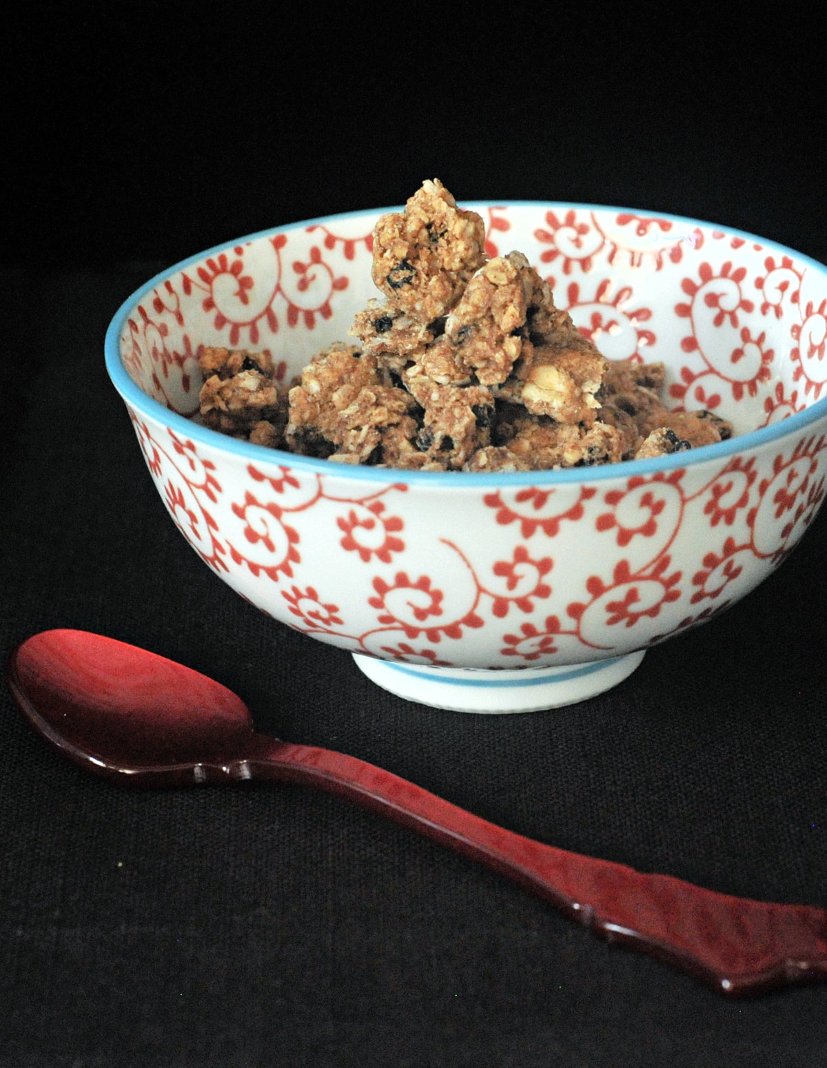 Oatmeal raisin cookie granola in a red and white cereal bowl, a red spoon next to bowl.