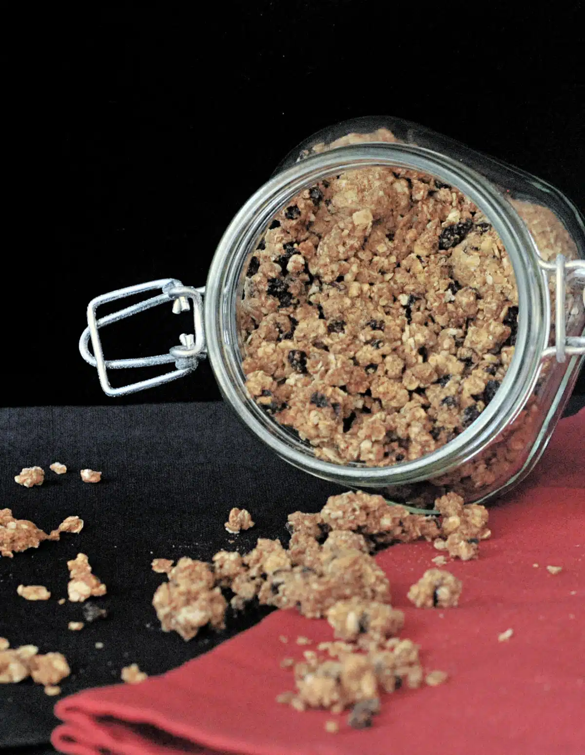 Oatmeal raisin cookie granola spilling out of a glass jar onto a red napkin on a black surface.