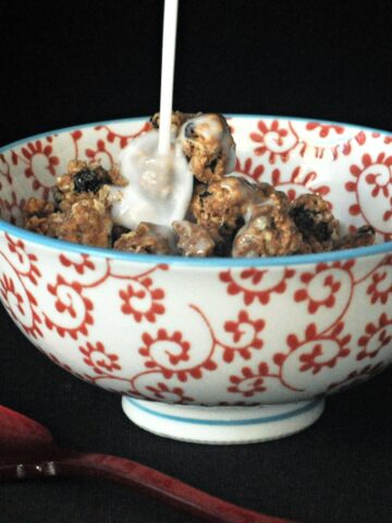 Milk being poured over oatmeal raisin cookie granola in a red and white cereal bowl, a red spoon next to bowl.