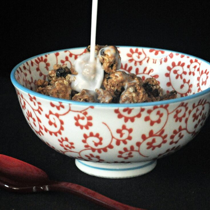 Milk being poured over oatmeal raisin cookie granola in a red and white cereal bowl, a red spoon next to bowl.