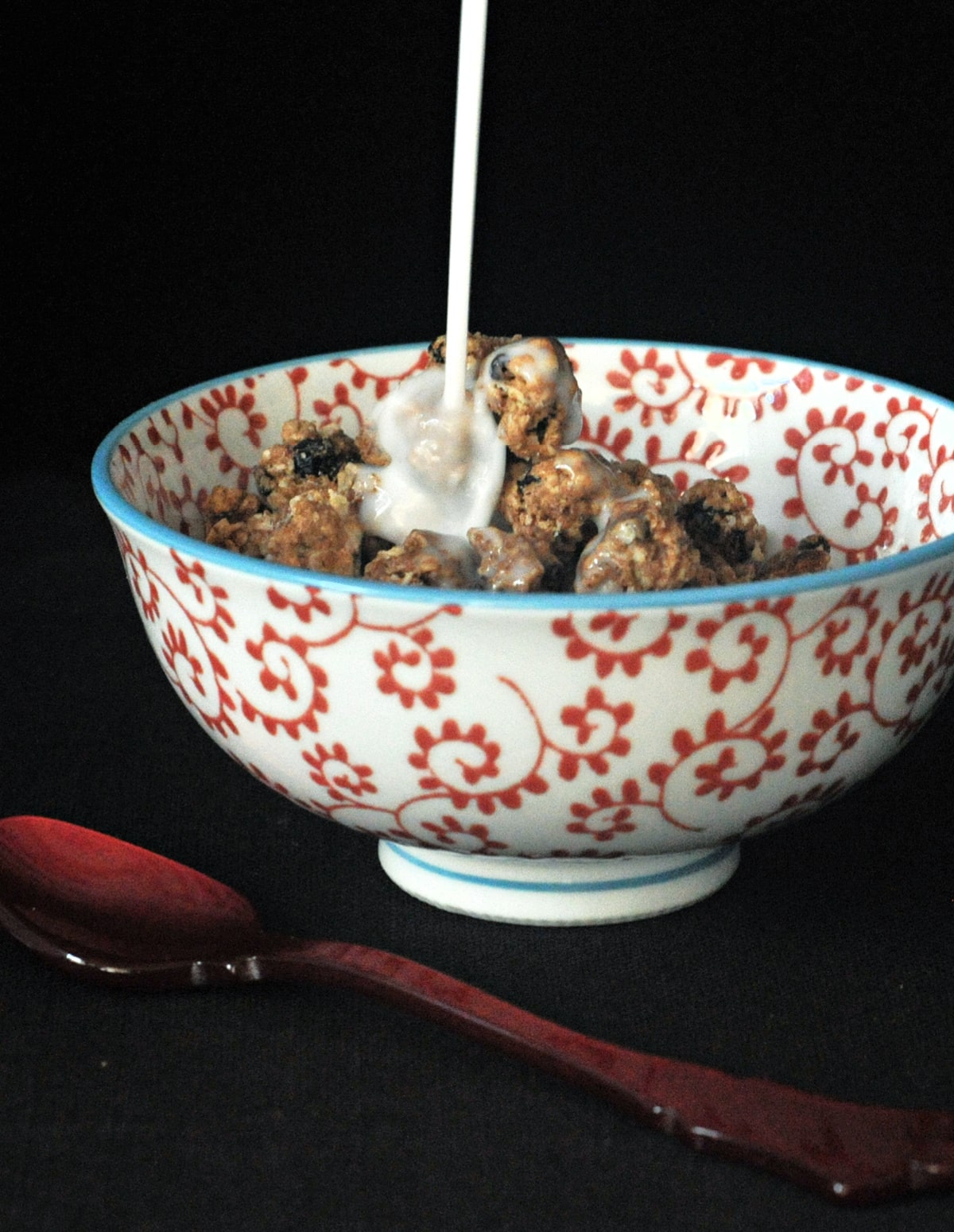 Milk being poured over oatmeal raisin cookie granola in a red and white cereal bowl, a red spoon next to bowl.