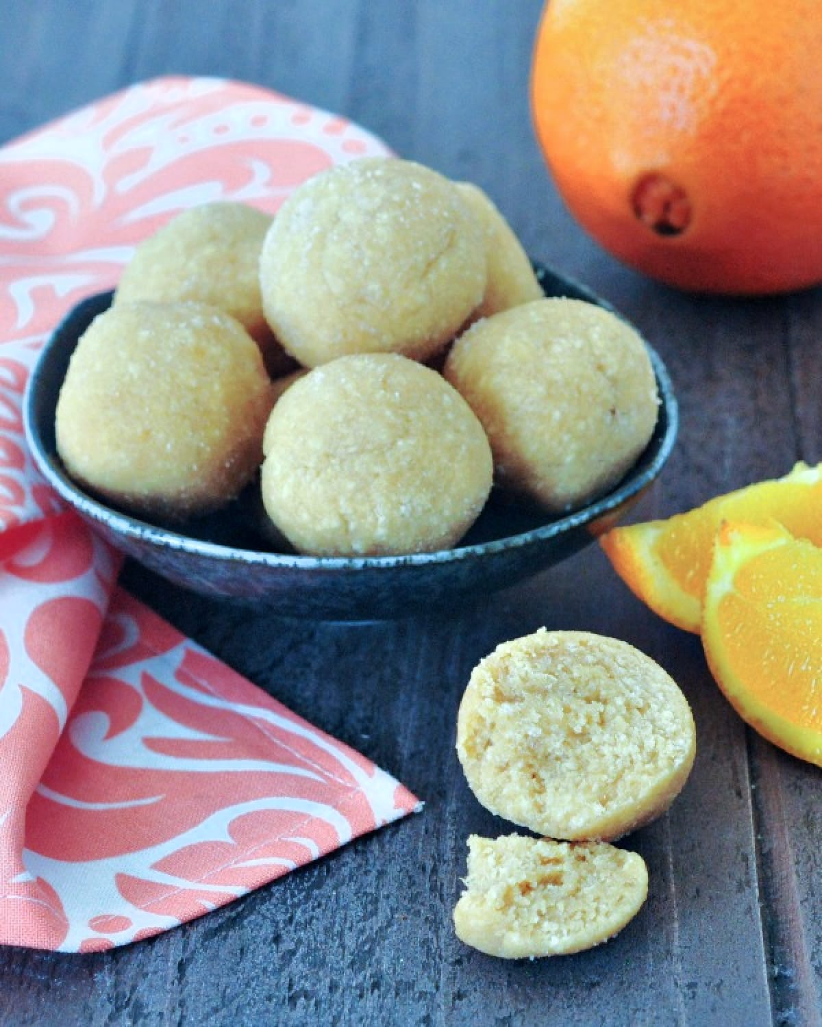 Small round orange creamsicle protein bites stacked in a rustic dark grey bowl, one bite sitting outside the bowl on a wood surface. Whole unpeeled orange and several fresh orange slices in background.