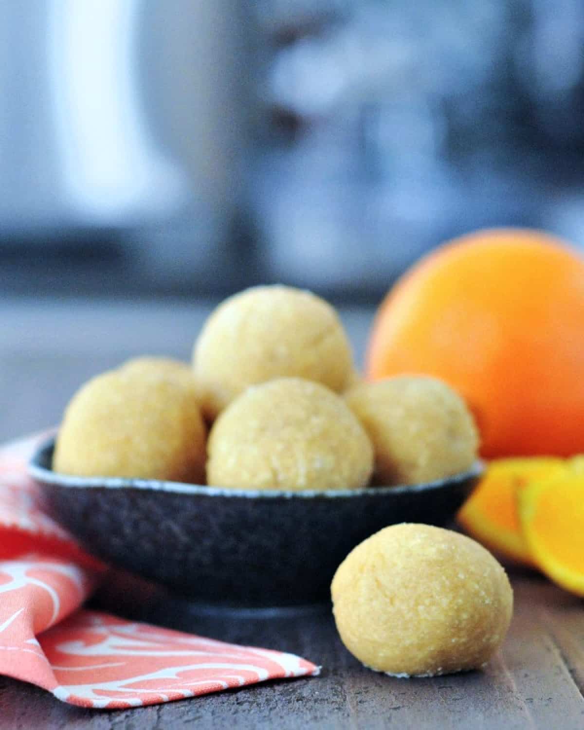 Small round orange creamsicle protein bites stacked in a rustic dark grey bowl, one bite sitting outside the bowl on a wood surface. Whole unpeeled orange and several fresh orange slices blurred in background.