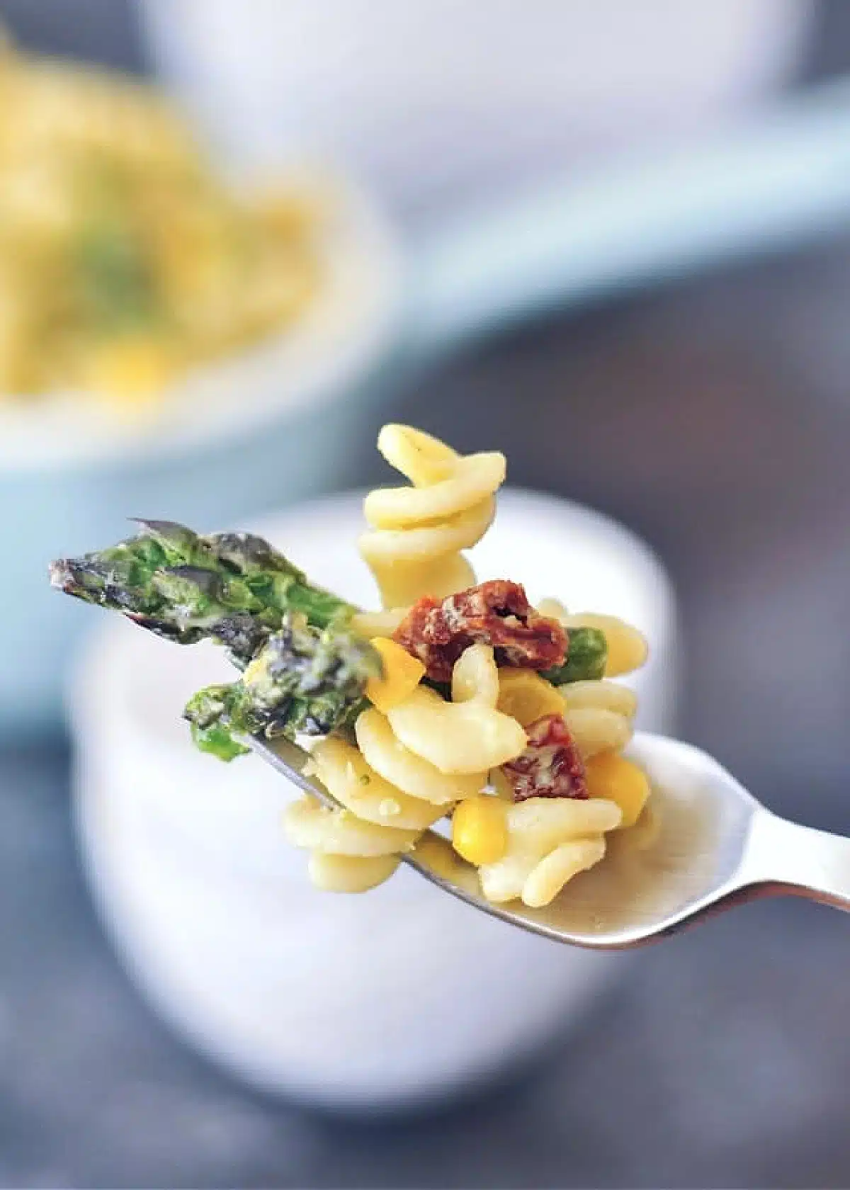 One single bite of picnic pasta salad on a fork: three corkscrew pasta noodles, two spears asparagus, several kernels corn, sundried tomato