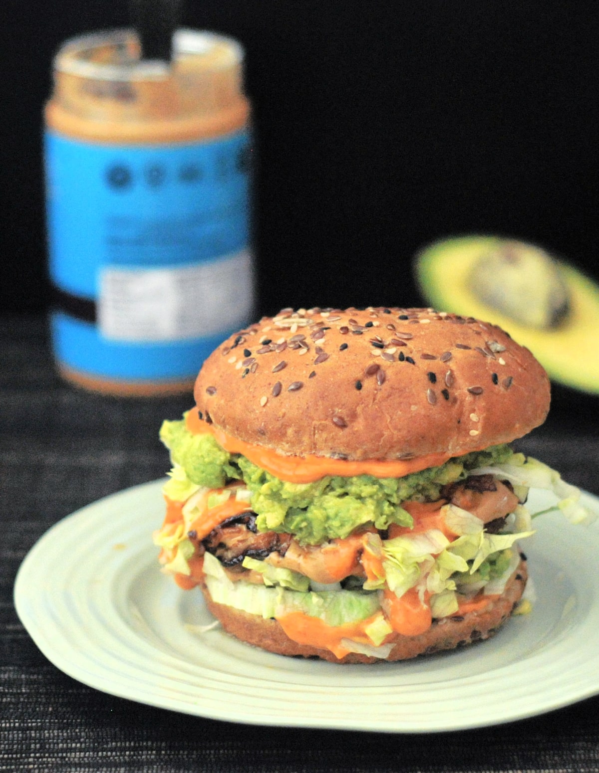 a spicy peanut butter burger on a light green plate against a black background; burger has bright orange spicy kimchi sauce, shredded iceberg lettuce, a veggie patty, mashed avocado, and a seeded bun. a jar of peanut butter and half an avocado blurred in background.