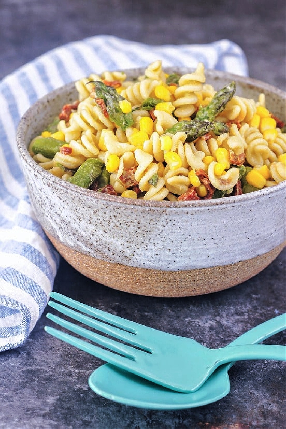 Picnic pasta salad in a large rustic bowl with bright blue serving utensils: curly pasta, asparagus, corn, sundried tomatoes