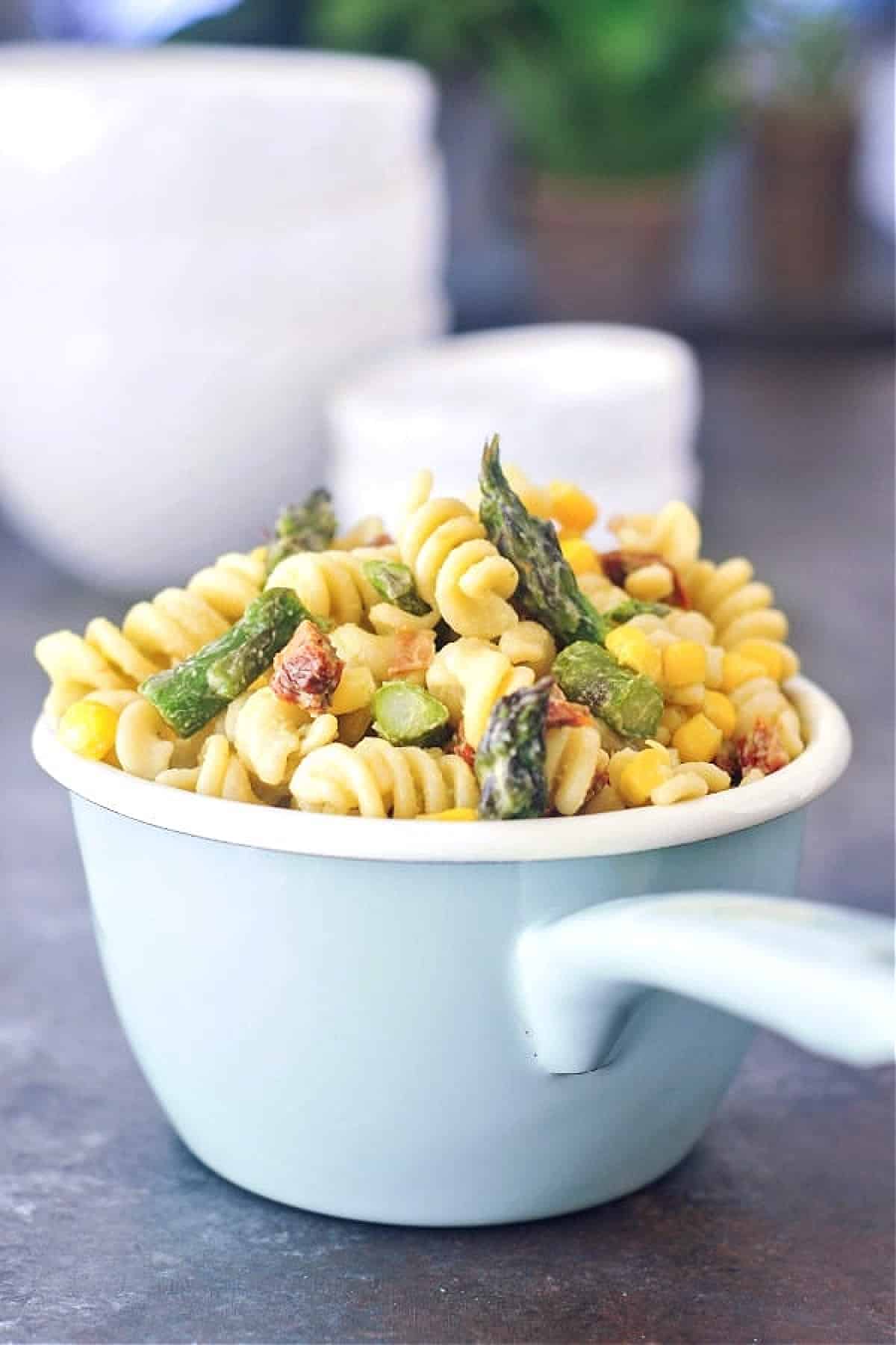 Vegan pasta salad (corkscrew pasta with sliced asparagus, fresh corn kernels, sundried tomatoes, avocado dressing) in a light blue long handled serving bowl.