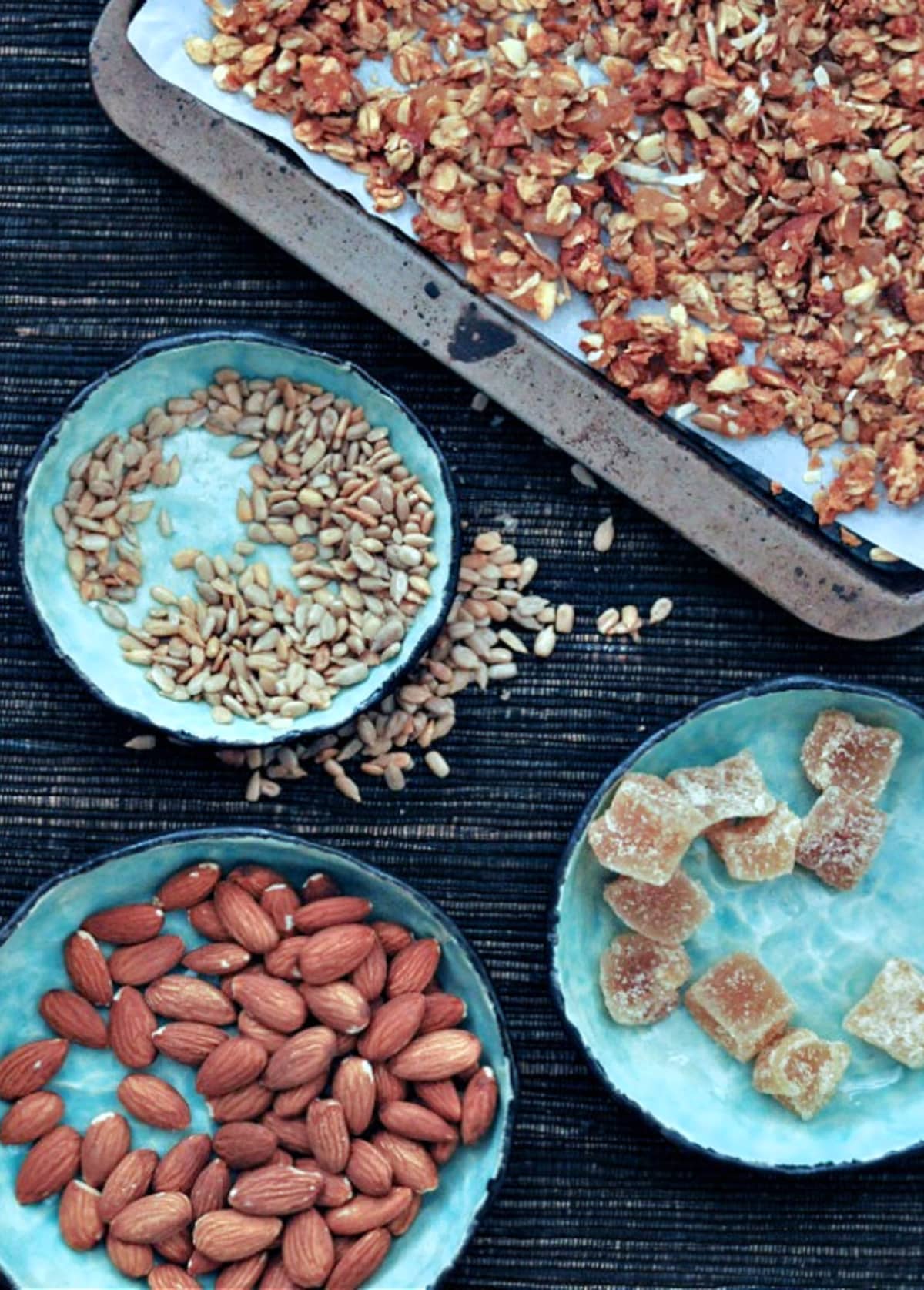 Overhead view of golden colored ginger vanilla granola on a parchment lined baking sheet. Bowls of the individual ingredients next to baking sheet: crystallized ginger, sunflower seeds, almonds.