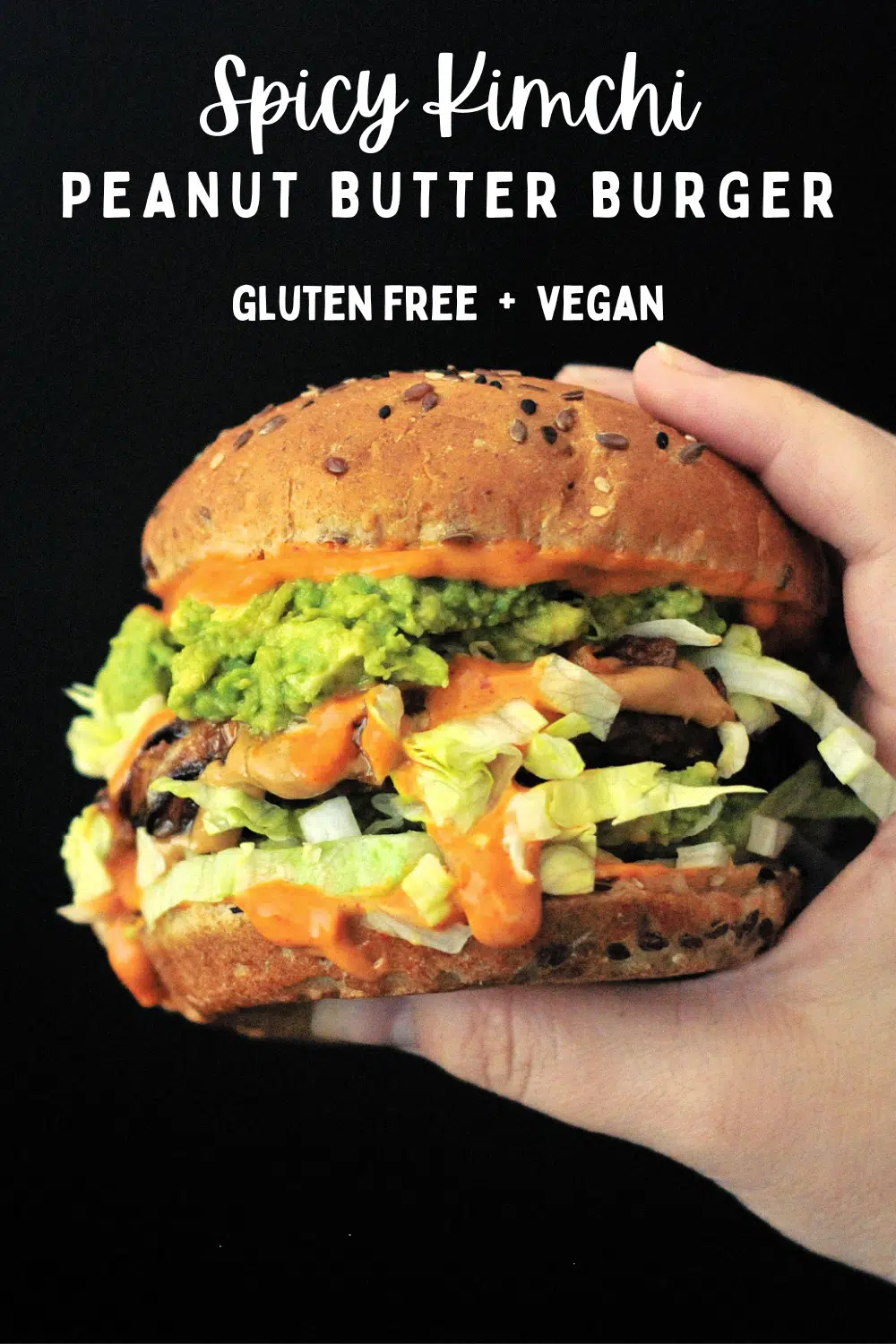 a spicy peanut butter burger held in a hand against a black background; burger has bright orange spicy kimchi sauce, shredded iceberg lettuce, a veggie patty, mashed avocado, and a seeded bun.