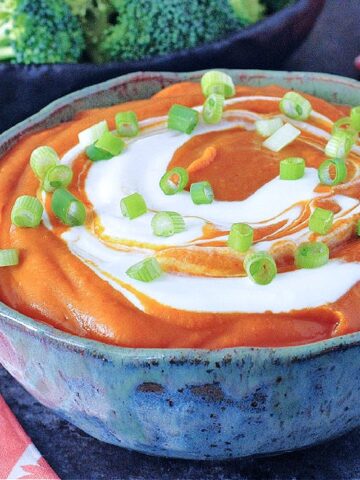 Deep orange enchilada dip with bright white sour cream swirled on top, garnished with chopped green onion. Dip is in a grey bowl with broccoli trees on the side for dipping.