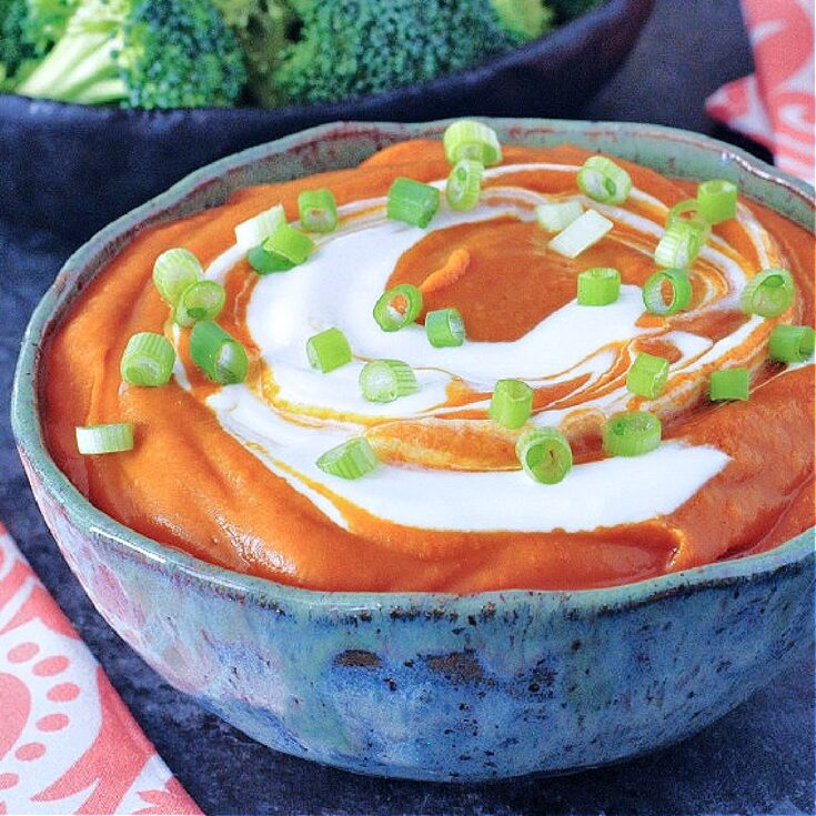Deep orange enchilada dip with bright white sour cream swirled on top, garnished with chopped green onion. Dip is in a grey bowl with broccoli trees on the side for dipping.