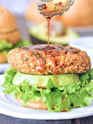 A ginger sesame teriyaki burger sitting on a bottom bun with lettuce and a thick slice of avocado, with more teriyaki sauce being spooned on top of the burger. Top bun blurred in background with a half of avocado, more burgers and buns.