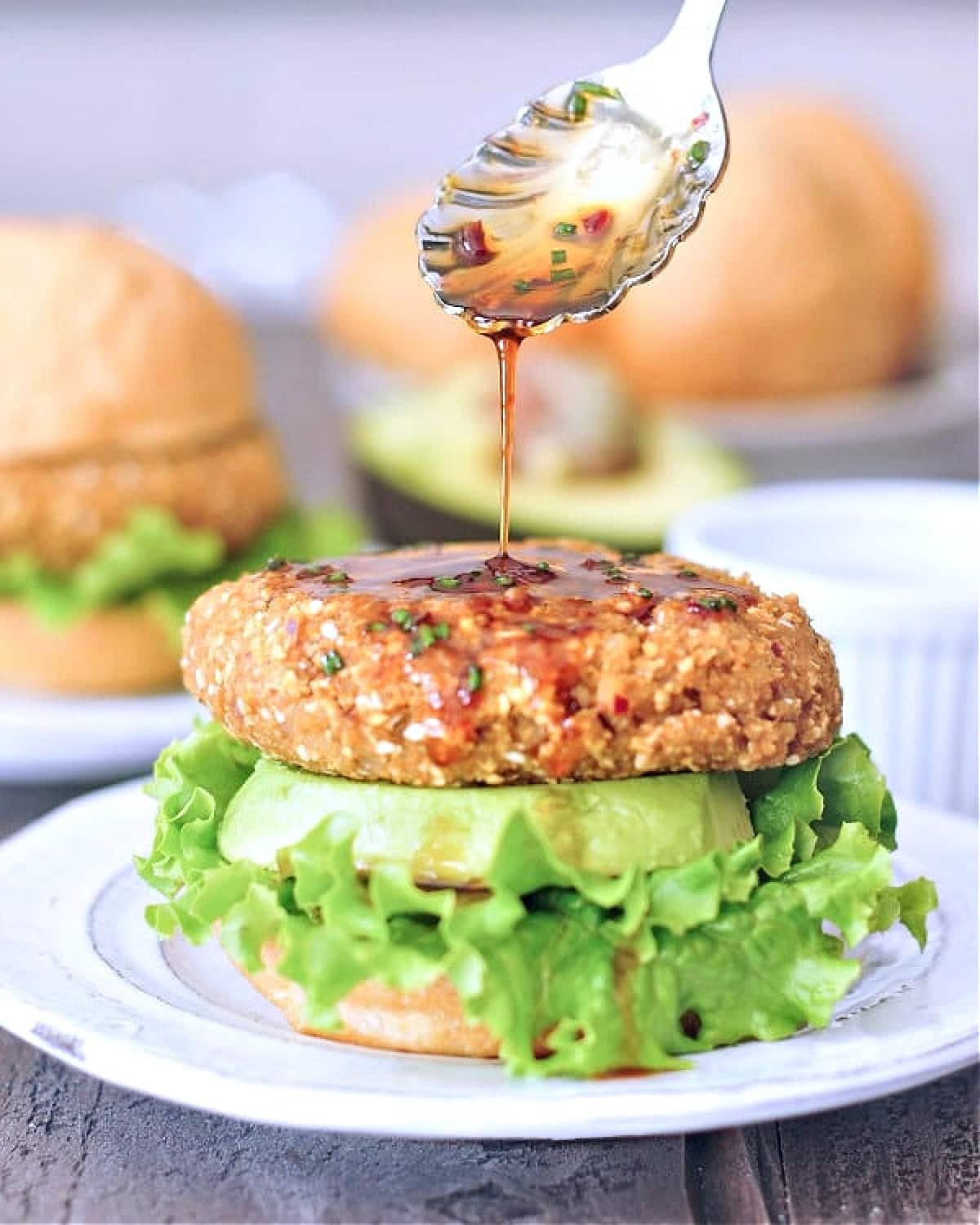 A ginger sesame teriyaki burger sitting on a bottom bun with lettuce and a thick slice of avocado, with more teriyaki sauce being spooned on top of the burger. Top bun blurred in background with a half of avocado, more burgers and buns.