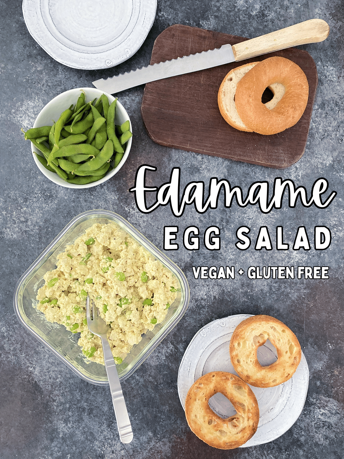 Overhead view of vegan edamame egg salad in a square glass container, with a small bowl of unshelled edamame, a small cutting board with a bagel and knife, and a plate with a toasted sliced bagel.
