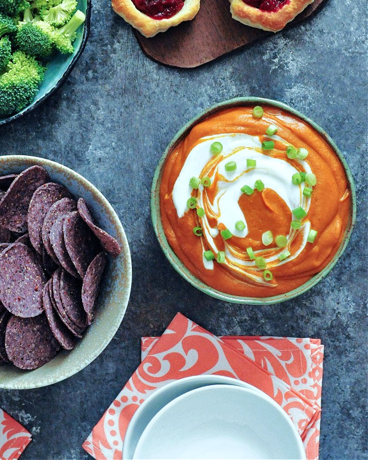 Overhead view of deep orange enchilada dip with bright white sour cream swirled on top, garnished with chopped green onion. Dip is in a grey bowl with broccoli trees and blue corn chips on the side for dipping.