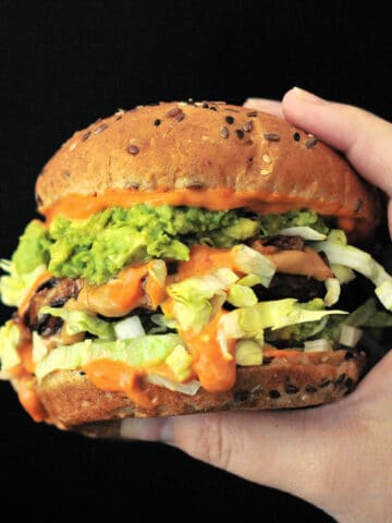 a spicy peanut butter burger held in a hand against a black background; burger has bright orange spicy kimchi sauce, shredded iceberg lettuce, a veggie patty, mashed avocado, and a seeded bun.