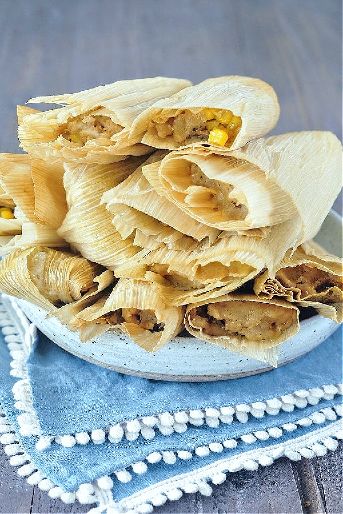 A plate of steamed vegan tamales wrapped in corn husks, stacked high in a shallow bowl sitting on a blue cloth napkin.