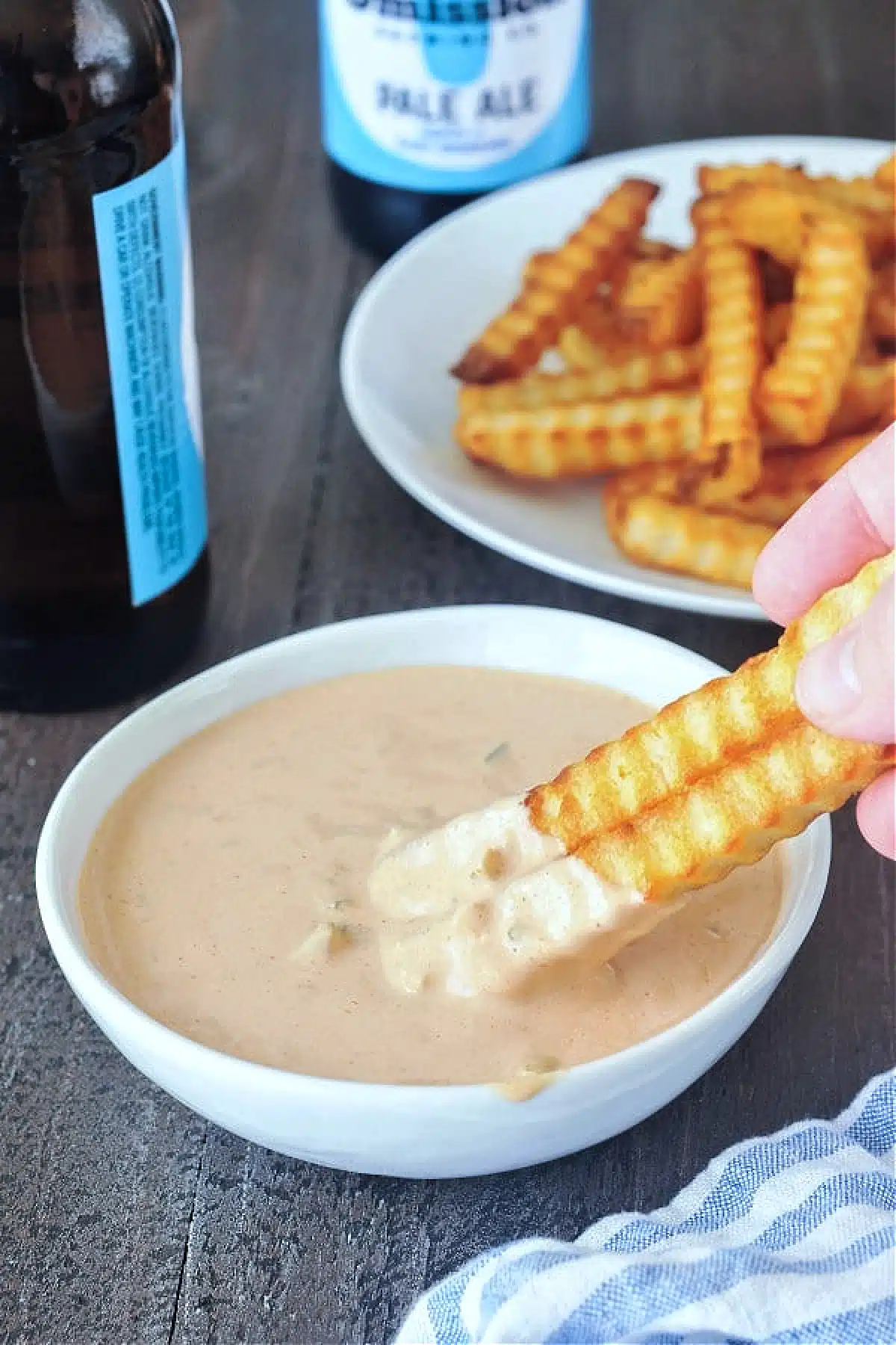 Krinkle cut French fries being dipped into a bowl of Thousand Island dressing, plate of fries and bottles of beer in background.