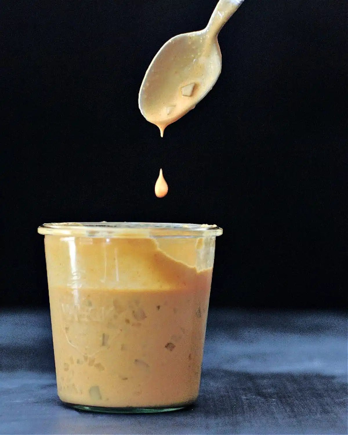 A large glass Weck jar of vegan thousand island dressing, with a spoon dripping sauce above the jar against a black background.