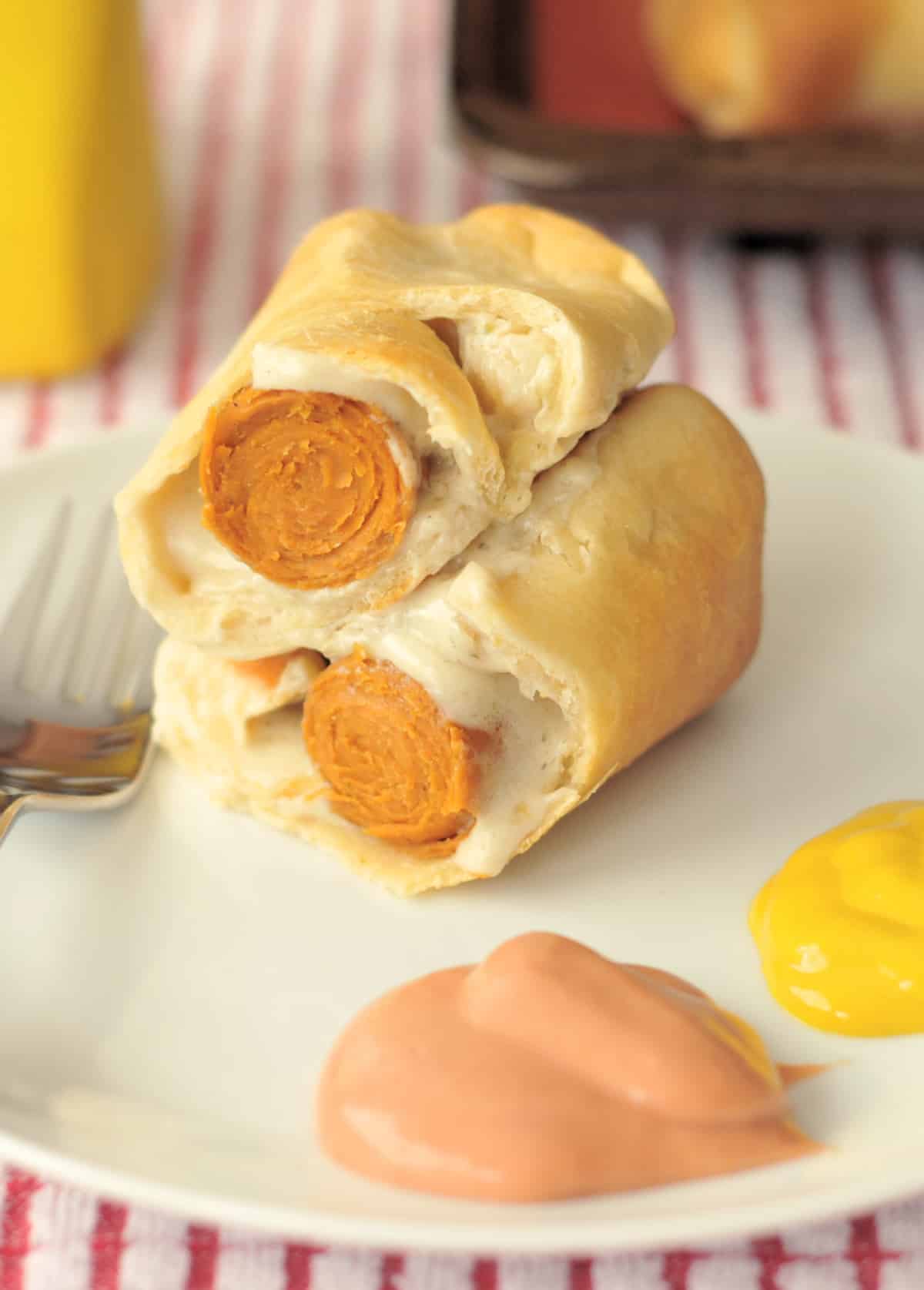 A hot dog with cheese wrapped in baked pastry cut in half and sitting stacked on a white plate, with fry sauce (ketchup and mayonnaise) and mustard. Bright yellow mustard bottle blurred in background.