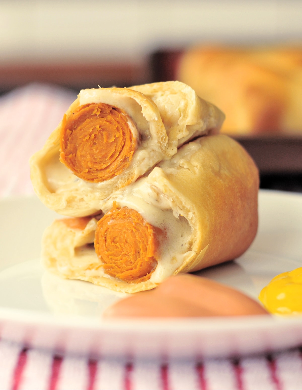 One wiener wrap (a hot dog with cheese wrapped in pastry and baked) cut in half and sitting stacked on a white plate, with fry sauce (ketchup and mayonnaise) and mustard.