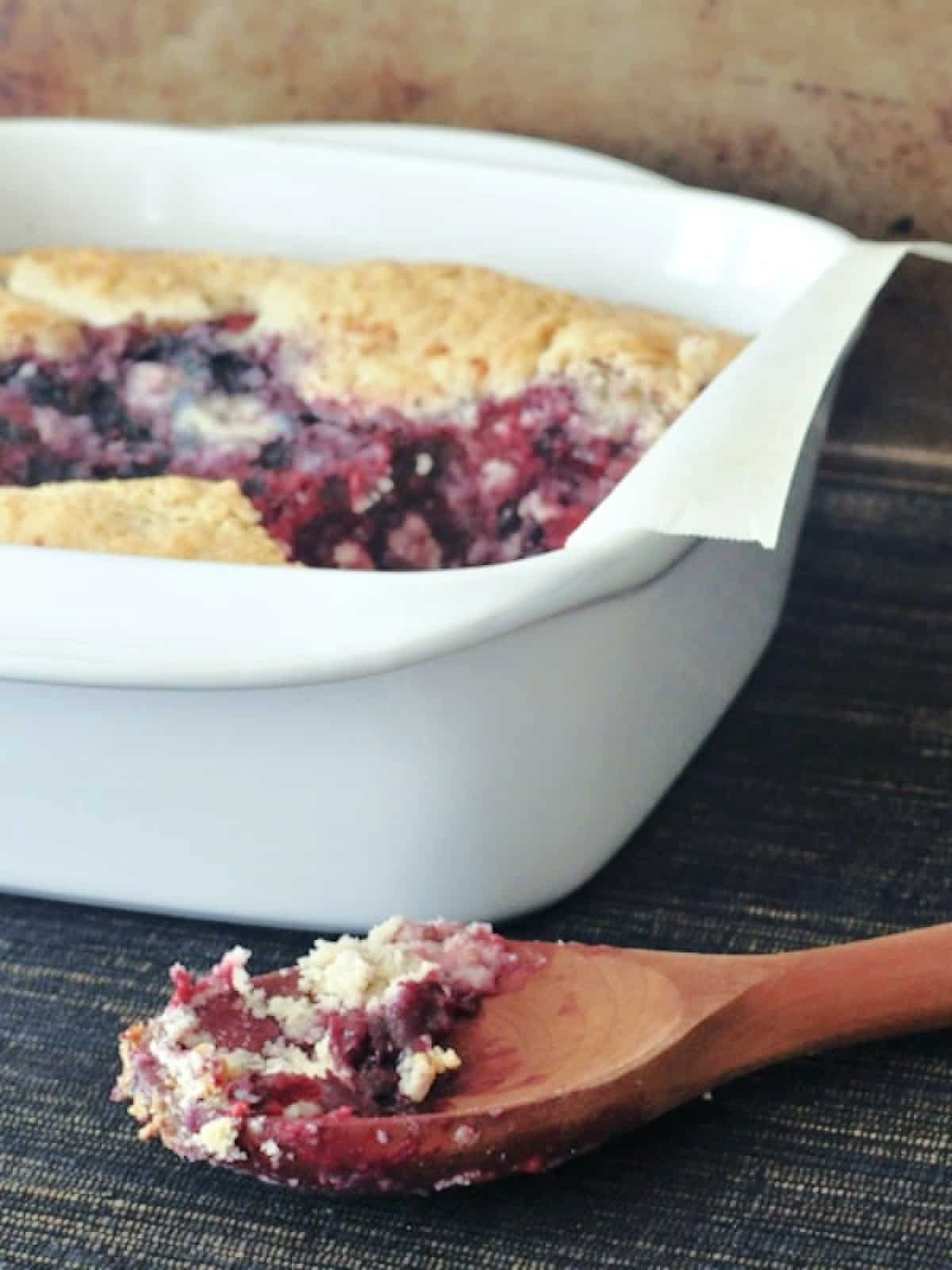 White square baking dish of mixed berry cobbler with a wooden spoon next to it with cobbler on the spoon.