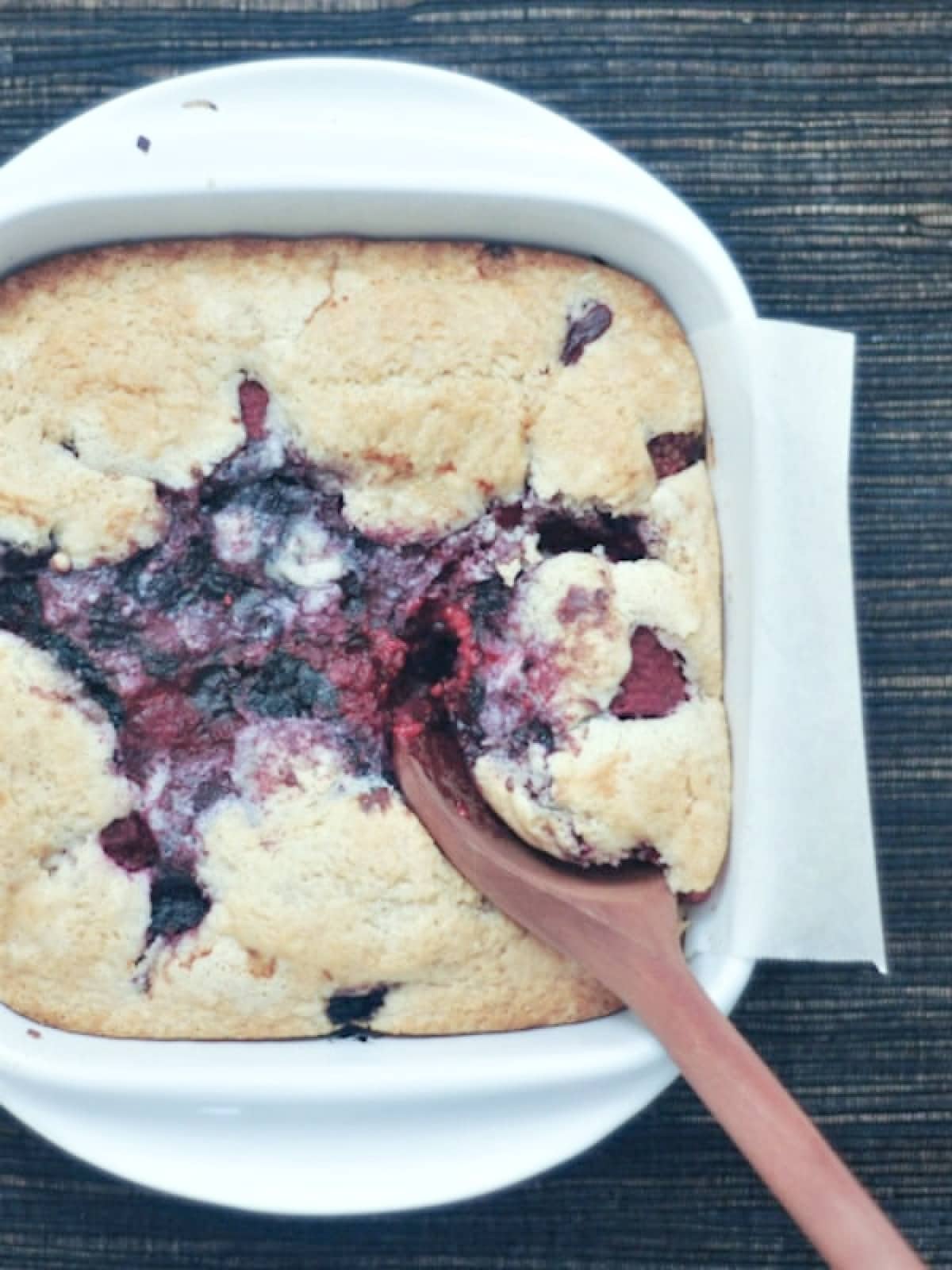 Overhead view of white square baking dish of mixed berry cobbler with a wooden spoon digging into it.
