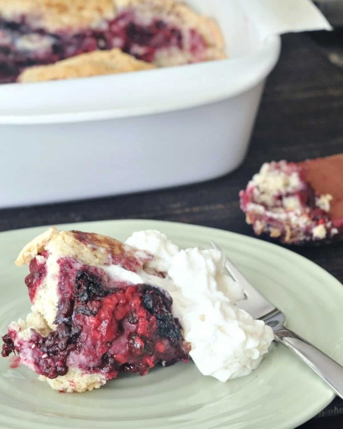 One serving of mixed berry cobbler on a plate, topped with whip cream Full baking dish of cobbler in background.