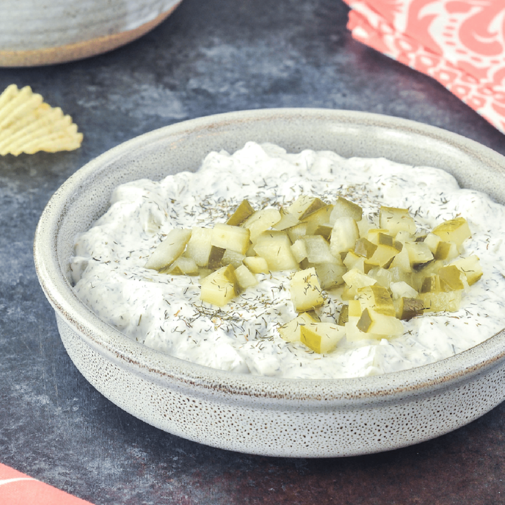 Dill pickle dip in a shallow bowl, with a second bowl of ruffled potato chips for dipping.